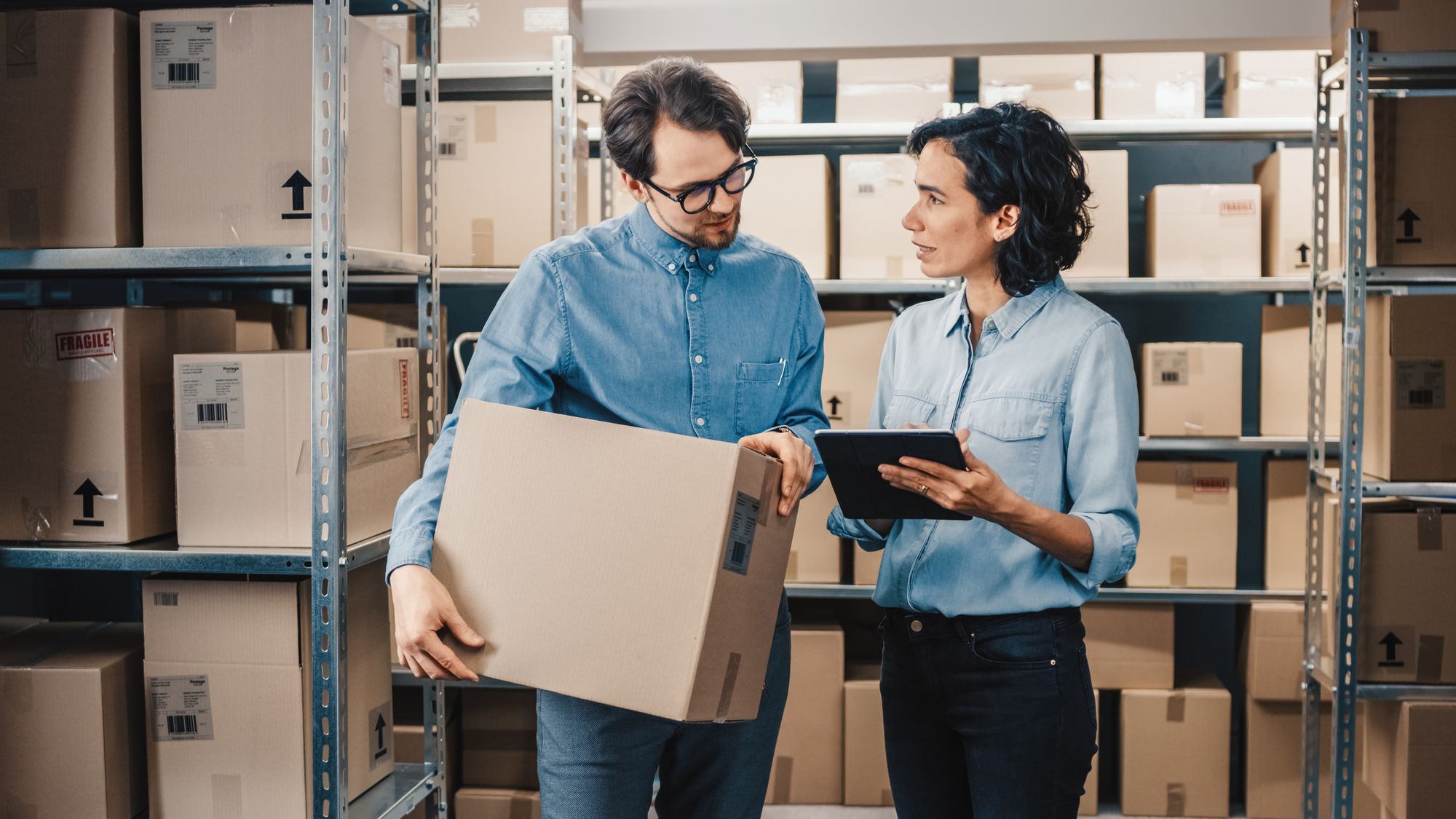 A male employee who holds a boxful of inventory and a female employee checking inventory level on a tablet