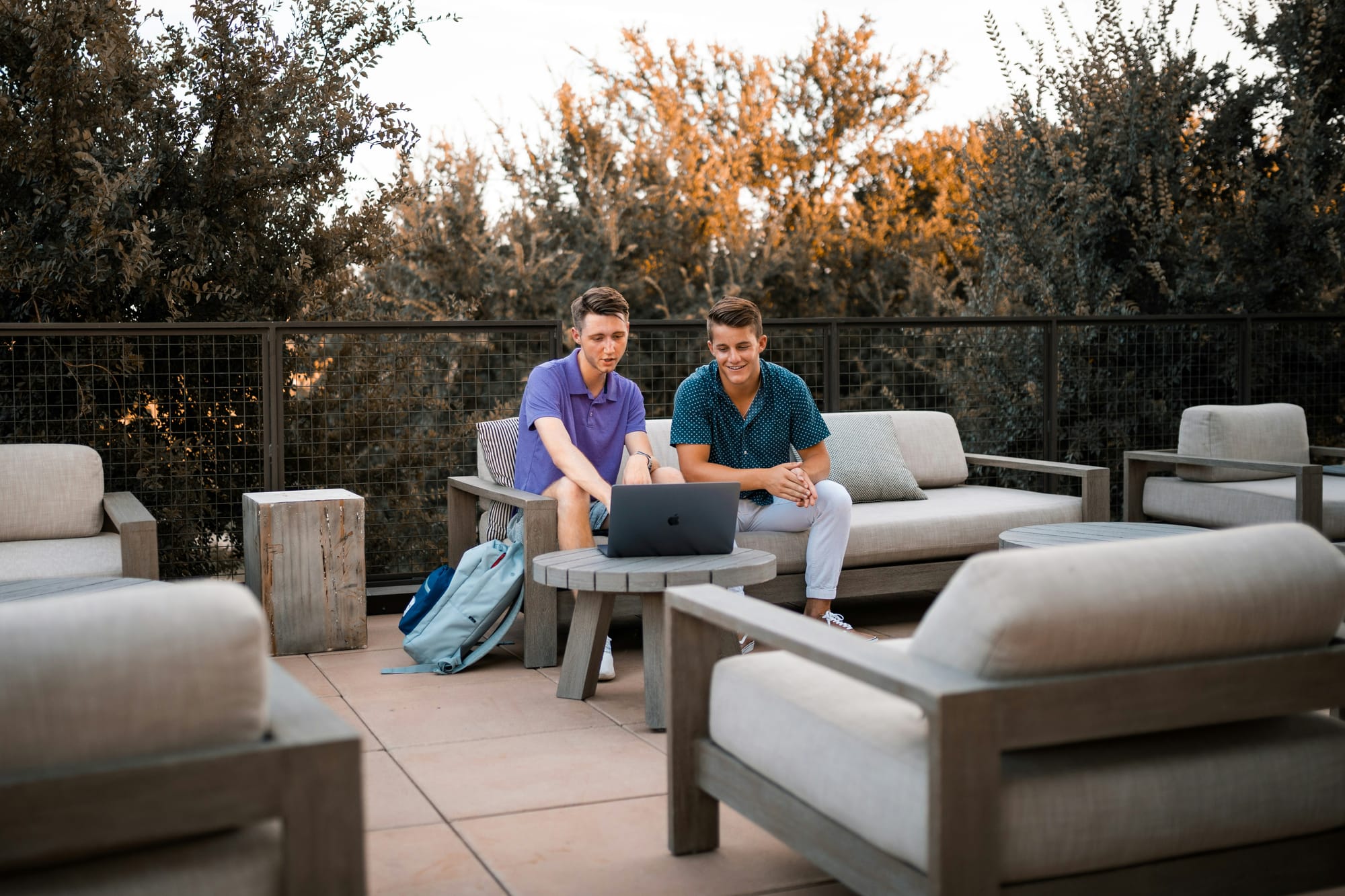 Two young men sitting outdoors on a patio, looking at a laptop together on a wooden table, surrounded by modern outdoor furniture.